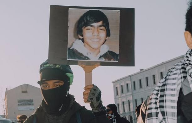 Vermummter Demonstrant*in mit einem Bild von Berkin Elvan, 2013 bei den Gezi-Protesten durch eine von der Polizei zur Schusswaffe umfunktionierten Tränengaskartusche ermordet. Von: cidemi.art